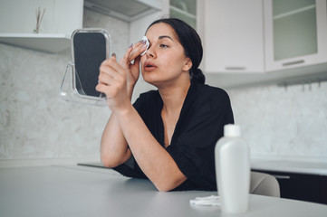 Obraz premium Beautiful brunette woman removing makeup from her face with mirror. Cute beautiful natural beauty girl cleaning face with cotton sponge. Cosmetology and spa, problem skin care, acne treatment concept