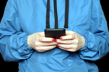 Young girl wearing in blue windbreaker looks to the screen of black smartphone photo on black background front close up © DyMaxFoto