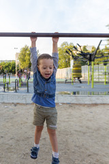 Fototapeta premium Happy small boy hanging on brachiating bar at a school yard. Other kids playing on levers for training in background. Selective focus.