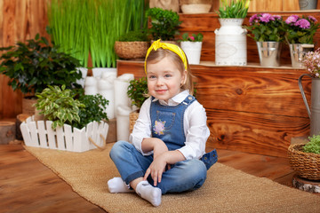 Smiling little girl with bandage her hair sits on wooden floor around green houseplants and flowers. Gardening. Child playing in backyard. Рappy child girl playning on terrace. Girl in kindergarten	
