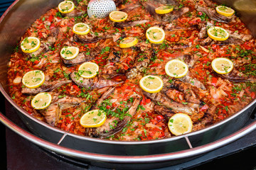 Close up of large portion of cooked fish with red peppers, onion, parsley and fresh yellow lemons in a large pan at a street food festival, ready to eat healthy seafood
