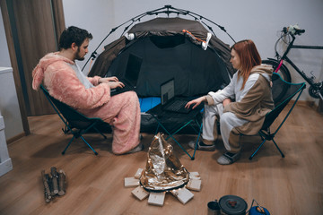 Couple sitting in folding chairs by the tent and bonfire. Camping in the apartment. Use laptop, notebook, work online. Remote job. Stay at home. Communications and Internet. quarantine.