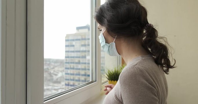Young woman in protective medical mask goes to the window in isolation for virus. Girl looks out the window during coronavirus pandemic. Health care and medical concept