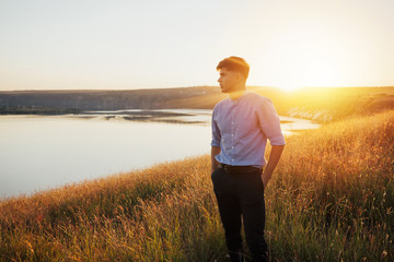 Free man in stylish classical clothes on sunset admire nature view of summer lake. Success. Travel. Good life.  Handsome man standing with hands on hips on hill and looking at lake. Copy space.