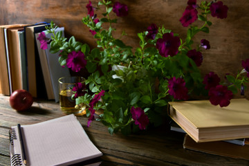 The photo shows an open notebook, a cup, an apple and books on a wooden table with flowers