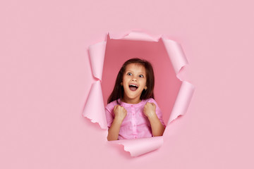 Portrait of cute happy smiling caucasian little child girl raises clenched fists and poses in blue paper hole.