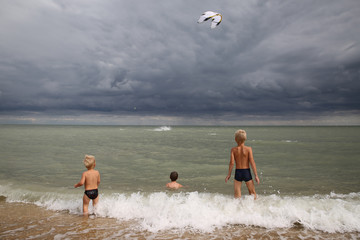 three boys stand in the waves and look at a kite in a stormy sky