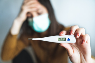 Selective focus of a sick young woman wearing a surgical mask and holding a digital thermometer that indicates she has over 38 degrees fever. Sick and worried female with fever and illness