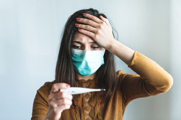 Sick young woman wearing a surgical mask and holding a digital thermometer that indicates she has...