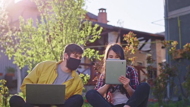 Young couple stay at home in the garden they take a sit on the grass working online while stay at quarantine they wearing protective masks using tablet and laptop to work lady showed something to her