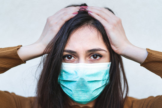 Close up portrait of disturbed annoyed young woman holding hands above head wearing a surgical mask feeling sick and worried about epidemic outbreak. Anxious lady touching head with stress migraine 