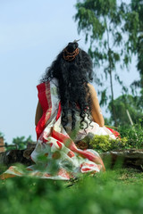 back side of a young lady in a hindu goddess devi outfit sitting in front of nature wearing red and white saree with long hair