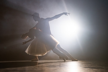 Graceful ballerina and her male partner dancing elements of classical or modern ballet in dark with floodlight backlight. Couple in smoke on black background. Art concept. © kohanova1991