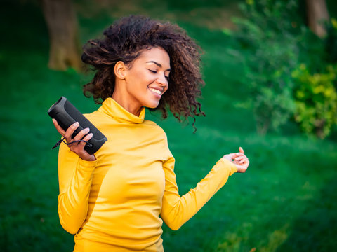Modern trendy african american girl listening to music by wireless portable speaker.Young beautiful woman enjoying, dancing in park.