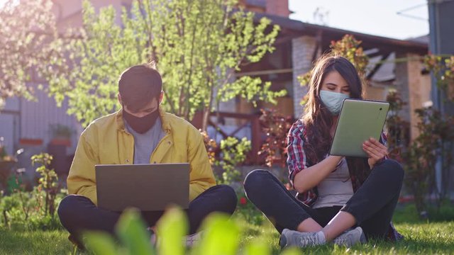 Stay at home in the garden sitting down on the grass concentrated lady and guy studying from home they wearing protective mask using laptop and tablet they are in quarantine Coronavirus mers 2019