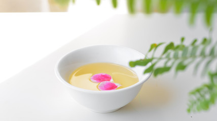 Fototapeta na wymiar Natural oil for beauty cosmetic procedures in a white bowl with pink petals on a white table against the background of tropical leaves. The concept of natural beauty.