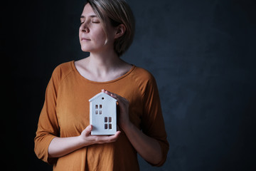 Horizontal shot of a young woman holding a ceramic toy cute house in her hands near the heart on a dark background. Empty space for text on the right. Caring for the house. Concept of stay at home