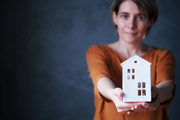 Horizontal shot of a young woman holding a ceramic toy cute house in her hands on a dark background. Focus in the foreground. Caring for the house. Concept of stay at home