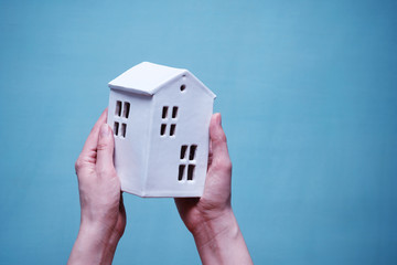 Hands of a young woman holding a small ceramic toy white house on a blue background. Horizontal shot with empty space at the top for text