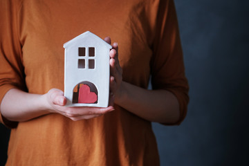 Horizontal shot of the hands of a young woman holding a ceramic toy cute home near a heart on a dark background. Empty space for text on the right. Caring for the house. Concept of stay at home