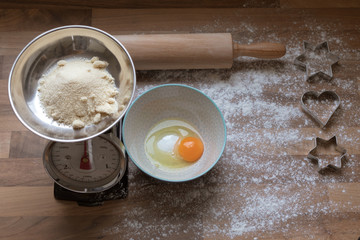 Top view of ingredients for preparation of homemade cookies showing an egg on vanilla sugar in a bowl, brown sugar on a scale, rolling pin and star and heart shaped cutting dies on a wooden table