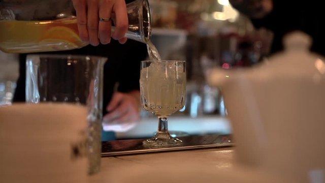 Bartender silhouette fills a transparent glass goblet with ice cubes and lemonade on a bar background, close-up.