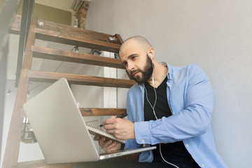 A guy in a blue shirt and black jeans is sitting on the stairs in the house and working at the computer. The concept of freelance work