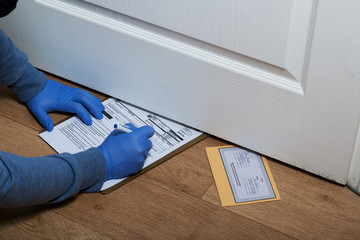 Delivery of correspondence during the period of self-isolation. A man in rubber gloves and a mask signs a letter delivery notification. It is inserted by the postman into the slot under the door.