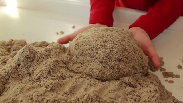 Brown kinetic sand quickly slides and falls from female hands, close-up. A teacher shows how to use an educational toy in a class filled with sunlight.