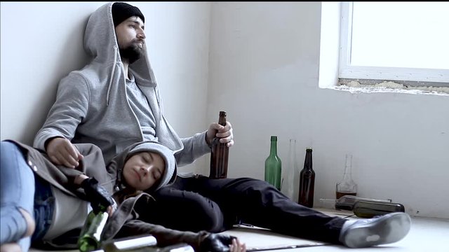 Homeless bums young man and woman are lying on cardboard on floor in abandoned building. Drunkard girl is sleeping and boy is drinking beer. Alcohol abuse addiction and street life concept.