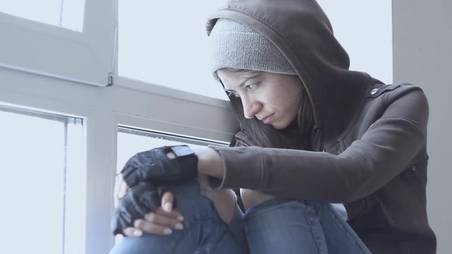 Lonely, depressed, upset young woman in hood is sitting on windowsill and crying in abandoned building. Homeless teenage girl ran away from home because of problems. Alcohol abuse, addiction concept.
