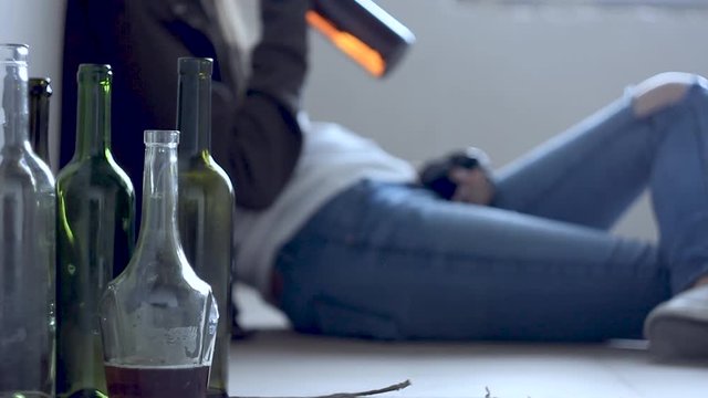Closeup empty glass bottles. On background homeless drunk woman is sitting on cardboard on floor in abandoned house. Addicted teenage girl drunkard is drinking beer. Alcoholism, alcohol abuse concept.