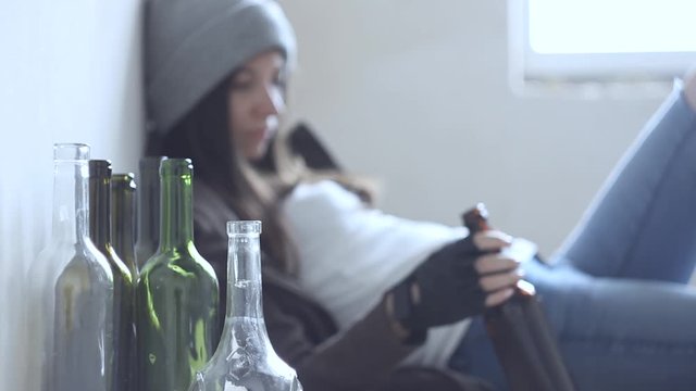 Closeup empty glass bottles. On background homeless drunk woman is sitting on cardboard on floor in abandoned house. Addicted teenage girl drunkard is drinking beer. Alcoholism, alcohol abuse concept.