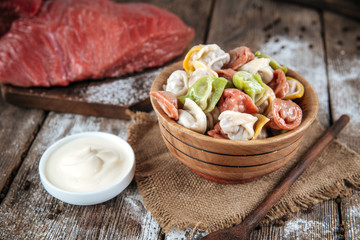 Side view on raw semi-finished multi-colored dumplings in a bowl on the wooden table with sour cream, horizontal