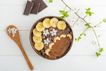 chocolate smoothie, banana with cashew in a coconut bowl, bamboo spoon on a white table with flowers. vegan breakfast and healthy food