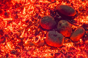 Potatoes in a peel on a blurry background of hot fiery heat. Top view on bright red embers and dark tubers of baked potatoes in a bonfire. Cooking in a barbecue outdoors. Close-up, selective focus
