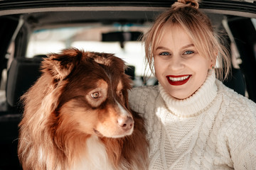 A beautiful woman is sitting and hugging, smiling in the trunk of a minivan with a dog breed Lassie Australian Shepherd. Caring for a dog. Dog is a mans friend, a true friend. Sunny winter day.