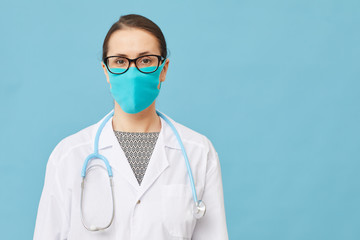 Portrait of young nurse wearing white coat and protective mask looking at camera over blue background
