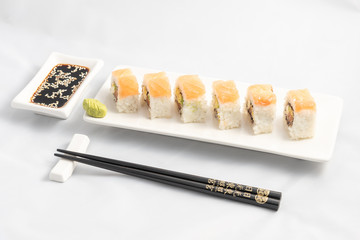 Sushi roll with pink salmon wrap, grilled salmon, Philadelphia cheese and avocado. White container and black chopsticks.