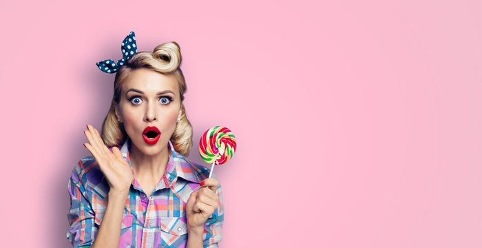 Excited surprised lovely woman with lollipop. Girl pin up with open mouth. Blond model at retro fashion and vintage concept. Pink color background. Copy space for some ad slogan, sign or text.