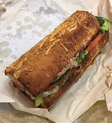 A toasted tuna fish grinder on herb and cheese bread unwrapped and sitting on the packaging 