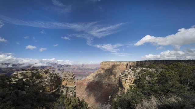 A wide motion timelapse shot of the Grand Canyon. The camera tracks left and tilts up to show more of the clouds flowing over Moran Point and white cliffs and vegetation in the area.