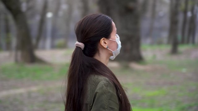 Lonely pretty young girl in protective medical face mask with brown ponytail long hair walks alone among trees in cold spring forest outdoor. Quarantine by reason of coronavirus covid-19 virus threat.