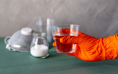 Hand in rubber orange color glove holding plastic cup with salty water to rinse nose and throat as prevention and protection from viruses. Covid-19 concept