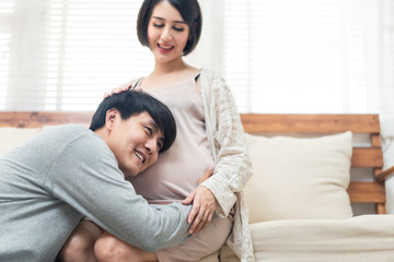 Asian man and pregnancy woman sit on sofa at home. Father holding mother tummy listen and hear infant inside mom belly. Husband sits and hugs wife. Love and care of pregnant and newborn kid concept.