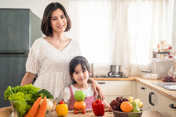 Lovely cute Asian family making food in kitchen at home. Portrait of smiling mother and daughter standing at cooking counter that food ingredient put on table. Happy family enjoy activity together.