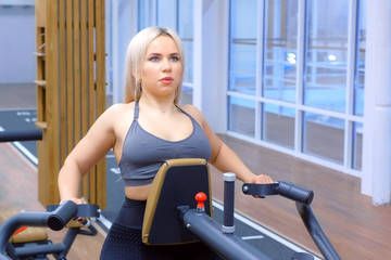 Fototapeta premium Sportive woman is doing exercises for spine muscles on block rowing machine. Sports workout in gym. Sport fitness concept. Blonde girl on persomal training making exercise for back.