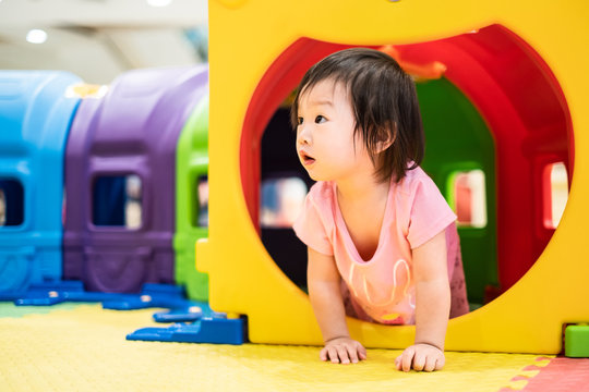 Little cute Asian toddler girl crawling out from colorful toy tunnel at playground. Kid enjoy exploring new things feeling fun and happy with smile. Learn & play education, development of child growth