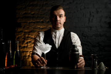 Barman holds transparent glass full of ice and shaker which stands on bar counter