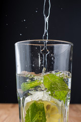 Close-up of glass with mint and lemon with water falling on wooden table and black background, vertical with copy space